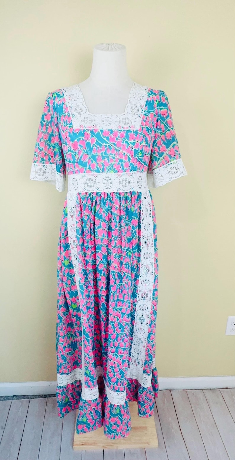 Pu&ograve; includere: Un abito lungo fino ai piedi con sfondo color ottanio e un motivo floreale rosa ripetuto. Il capo presenta bordi in pizzo bianco lungo lo scollo quadrato, i polsini delle maniche, il punto vita e i pannelli verticali della gonna.