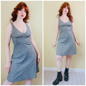 May include: A gray, sleeveless dress with a V-neckline and a subtle plaid pattern. The dress features a fitted bodice with two decorative buttons and a flared skirt. The dress is knee-length and appears to be made of a woven fabric.