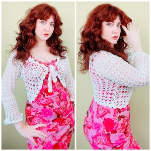 May include: A white long-sleeved crochet bolero jacket with an open-knit pattern and a front tie closure, worn over a sleeveless pink dress featuring a vibrant floral print with various shades of red, pink, and magenta roses and paisley patterns.