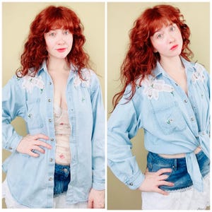 Può includere: Camicia di jeans azzurra con ricami floreali bianchi sulle spalle e piccoli accenti floreali verdi. La camicia ha due tasche frontali ed è abbottonata. Maniche lunghe con polsini abbottonati.