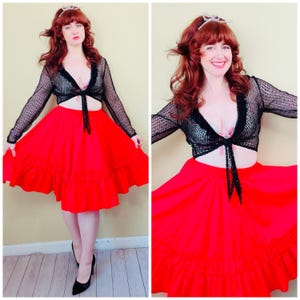 May include: A long-sleeved black mesh tie-front bolero top with a scalloped edge worn over a light-colored bralette, paired with a vibrant red knee-length skirt featuring a wide, gathered ruffle hemline and a full, flared silhouette.