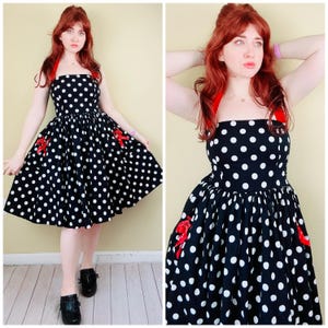 May include: A black knee-length dress featuring a white polka dot pattern, a straight neckline, and red ribbon accents on the bodice and pockets. The dress has a fitted bodice and a full, pleated skirt with a halter-style strap design.
