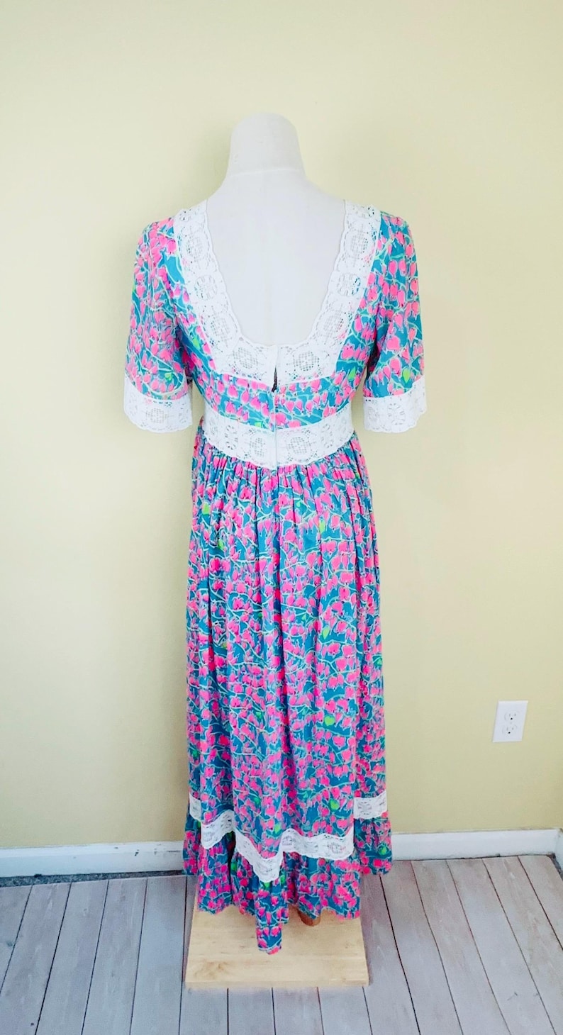 Pu&ograve; includere: Un abito lungo fino ai piedi con sfondo color ottanio e un motivo floreale rosa ripetuto. Il capo presenta bordi in pizzo bianco lungo lo scollo rotondo, la vita, i polsini delle maniche e una fascia orizzontale vicino all'orlo inferiore.