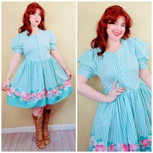 May include: A knee-length dress featuring a light blue and white vertical striped pattern with short puff sleeves and a button-down front. The hem is decorated with a vibrant floral print of pink and white roses and green leaves.