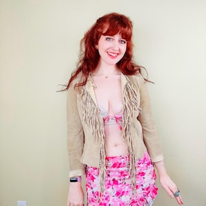 Könnte beinhalten: Eine beige Wildlederjacke mit Fransen, kombiniert mit einem pinkfarbenen Minirock mit Blumenmuster. Der Rock hat eine weiße Spitzenborte. Das Outfit wird durch Ringe und einen Fitnesstracker ergänzt. Der Stil ist bohème.