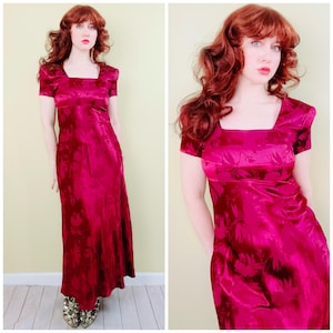 May include: A magenta-colored, floor-length dress with a square neckline and short sleeves. The dress has a subtle floral pattern and a fitted bodice. The model is wearing gold-colored shoes.
