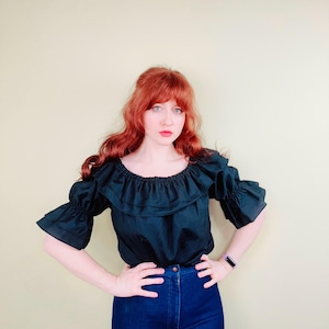 May include: A black off-the-shoulder top with ruffled neckline and sleeves. The top is paired with high-waisted, dark blue jeans. The model is standing in front of a light yellow background.