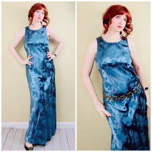 May include: A sleeveless, floor-length blue dress with a floral pattern. The dress has a scoop neckline and a fitted bodice. A gold-colored belt with heart-shaped links accents the waist. The dress is made of a shiny, silky material.