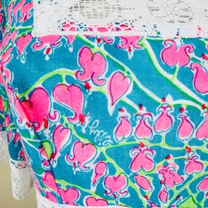 Pu&ograve; includere: Un abito turchese con un motivo ripetuto di fiori di cuore sanguinante rosa con steli verdi. Il capo presenta un bordo in pizzo bianco lungo lo scollo quadrato e la vita, con la firma Lilly Pulitzer stampata all'interno del disegno floreale.
