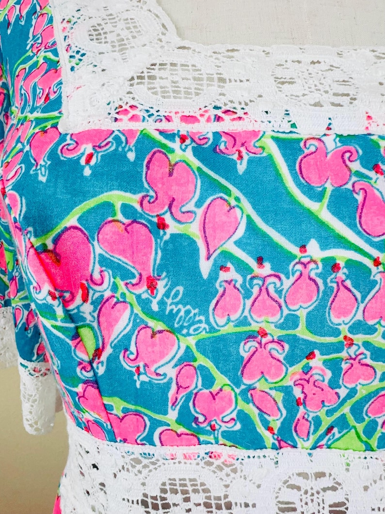 Pu&ograve; includere: Un abito turchese con un motivo ripetuto di fiori di cuore sanguinante rosa con steli verdi. Il capo presenta un bordo in pizzo bianco lungo lo scollo quadrato e la vita, con la firma Lilly Pulitzer stampata all'interno del disegno floreale.