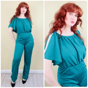 Könnte beinhalten: Türkisfarbener Jumpsuit mit Taillenbund und weitem Bein. Das Oberteil hat einen U-Boot-Ausschnitt und kurze, flatternde Ärmel. Der Jumpsuit ist aus einem glatten, einfarbigen Stoff gefertigt. Das Model trägt schwarze Schuhe.