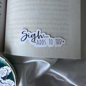 May include: A white sticker with the text "Sigh... *ADDS TO TBR*" in black script. The sticker is placed on the edge of a book. The sticker has a whimsical design, perfect for book lovers.