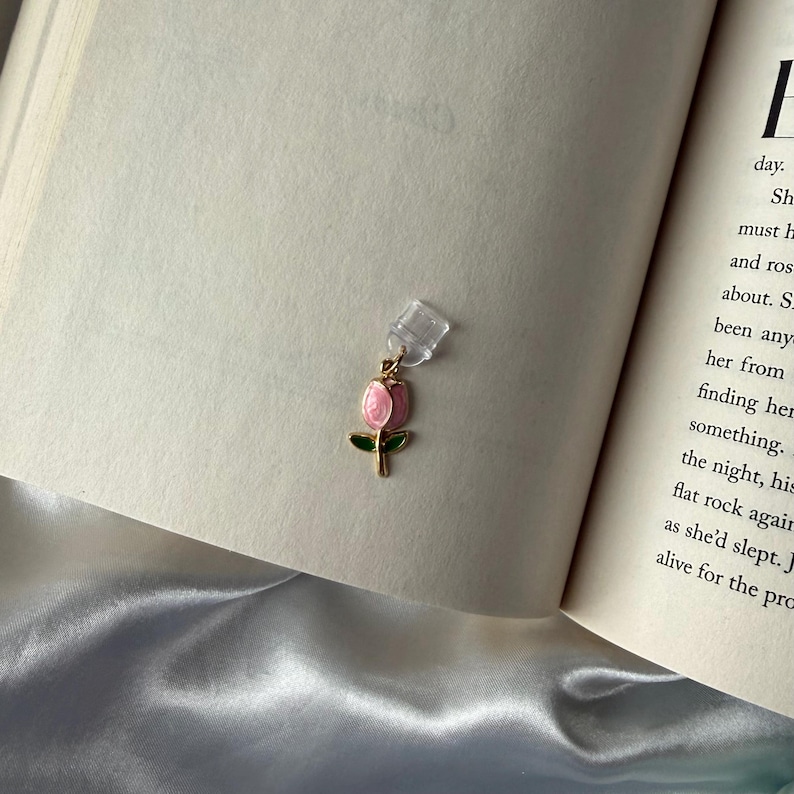May include: A gold-toned bookmark featuring a pink tulip design with green leaves. The bookmark is attached to a clear plastic clip, resting on the pages of an open book. The book's pages are cream-colored, and the background is a soft, white fabric.