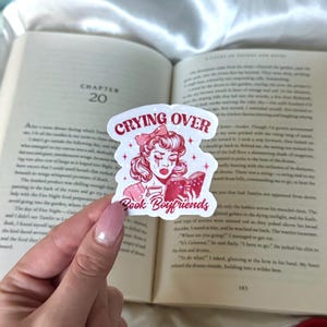 Puede incluir: Una pegatina roja y blanca con el texto "CRYING OVER Book Boyfriends" y una ilustración de una mujer leyendo un libro. La pegatina tiene un efecto brillante y se sostiene sobre un libro abierto.