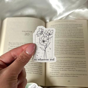 May include: A white sticker with a floral design and the text "to whatever end." The sticker features three flowers, a crescent moon, and stars within a rectangular frame. The sticker is held in front of an open book.