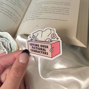 Puede incluir: Una pegatina con una caja de pañuelos rosa con el texto "Crying Over Fictional Characters". La pegatina tiene un diseño de pañuelo blanco y detalles florales. Sobre una superficie satinada con un libro abierto al fondo.