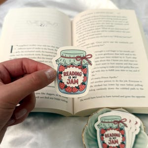 May include: A sticker featuring a jar filled with strawberries and the text "READING IS MY JAM." The jar has a light blue lid and a red bow. The sticker is held in front of an open book.