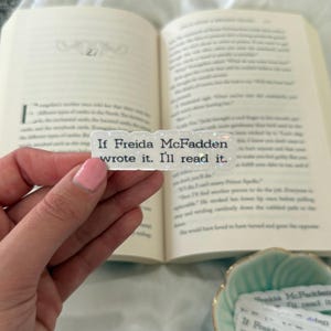 May include: A rectangular, glittery sticker with the text "If Freida McFadden wrote it. I'll read it." is held in front of an open book. The sticker has a white background with black text.