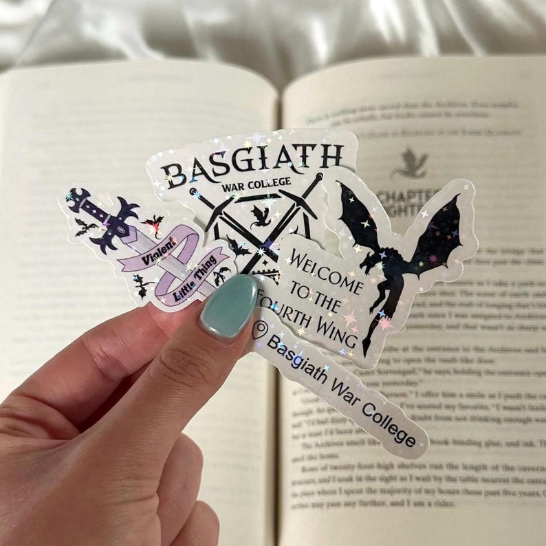 Peut inclure: Plusieurs autocollants holographiques avec des textes et des illustrations. On peut lire "BASGIATH WAR COLLEGE", "WELCOME TO THE FOURTH WING" et "Violent Little Thing". Les dessins incluent des &eacute;p&eacute;es, un dragon et un marqueur de localisation.