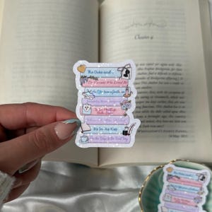 May include: A sticker featuring a stack of books with titles like "The Duke and" and "The Viscount Who Loved Me." The sticker has a glittery finish and includes small illustrations of a heart, a mask, and a quill.