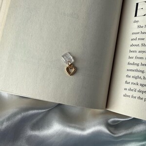 May include: A gold-colored heart-shaped charm with a textured surface, attached to a clear plastic clip. The charm is resting on the open pages of a book. The book's pages are off-white, and the background includes a satin fabric.