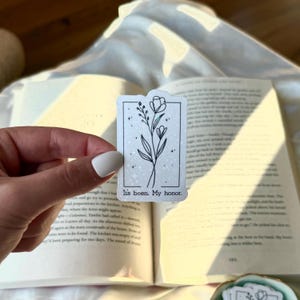May include: A rectangular sticker with a floral design and the text "It's been. My honor." The black and white illustration features a flower and leaves within a rectangular border. The sticker is held over an open book.