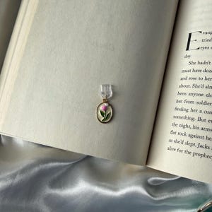 May include: A gold-toned bookmark with a pink and green floral design, attached to a clear plastic clip, rests on the open pages of a book. The bookmark's oval pendant features a delicate painted flower, adding a touch of elegance.