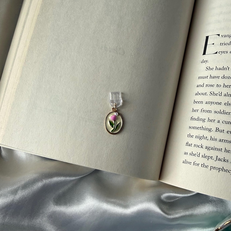 May include: A gold-toned bookmark with a pink and green floral design, attached to a clear plastic clip, rests on the open pages of a book. The bookmark's oval pendant features a delicate painted flower, adding a touch of elegance.