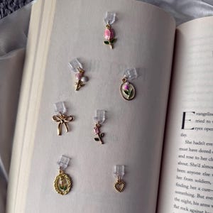 May include: A collection of gold-toned earrings with various designs, including a bow, a heart, and floral motifs. The earrings feature pink and green enamel details. The earrings are displayed on the pages of an open book.
