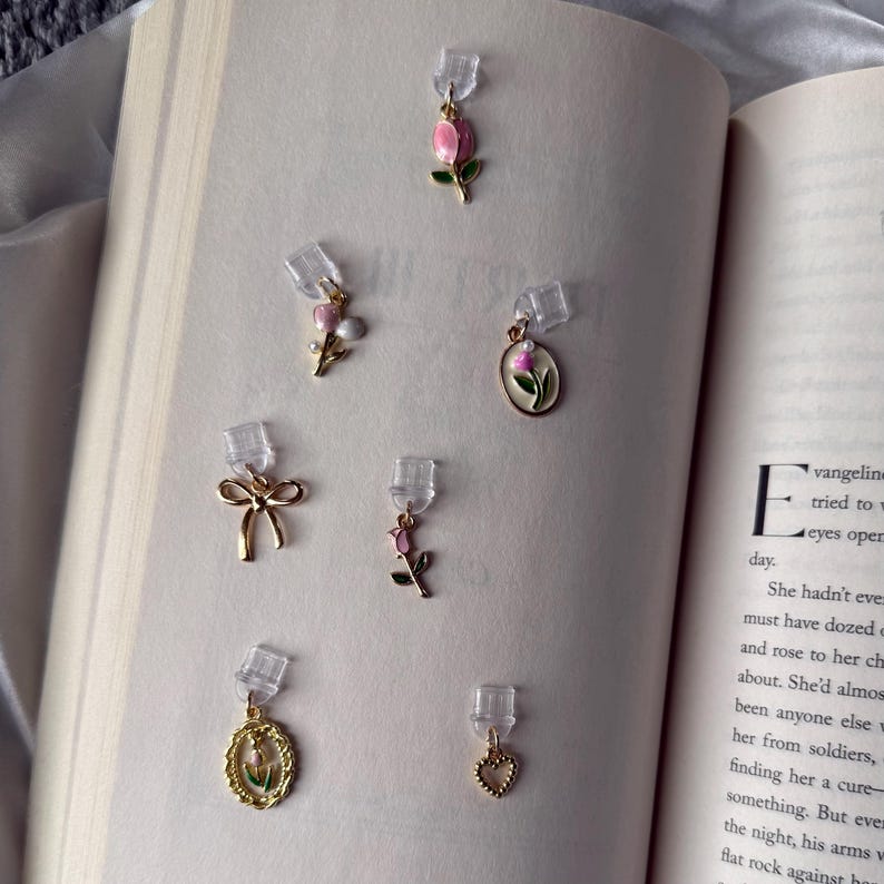 May include: A collection of gold-toned earrings with various designs, including a bow, a heart, and floral motifs. The earrings feature pink and green enamel details. The earrings are displayed on the pages of an open book.