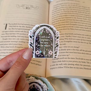 May include: A decorative sticker with a black arched frame design, featuring floral and star accents, and the text "I still read fairytales, but now for the villain." The sticker is held in front of an open book.