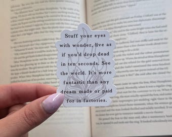 Puede incluir: Una pegatina blanca con la cita "Stuff your eyes with wonder, live as if you'd drop dead in ten seconds. See the world. It's more fantastic than any dream made or paid for in factories." La pegatina está delante de un libro abierto.