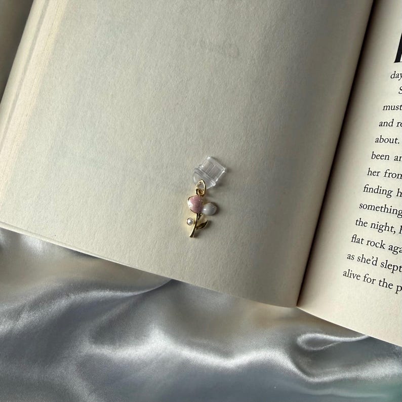 May include: A delicate gold-toned earring featuring a floral design with pink and white accents. The earring is adorned with small, sparkling details and a clear, square-shaped attachment. The earring is displayed on the pages of an open book.