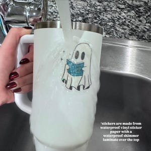May include: White tumbler with a silver rim and handle, featuring a ghost sticker reading a blue book. The sticker reads "A Court of Mist and Fury". Water streams over the tumbler. The text on the image says "stickers are made from waterproof vinyl sticker paper with a waterproof shimmer laminate over the top."