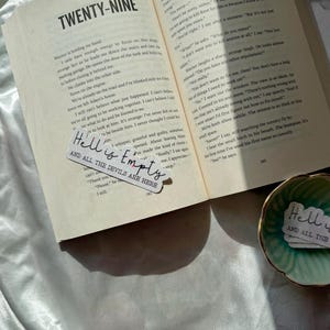 May include: An open book with the text "TWENTY-NINE" at the top. A sticker with the words "Hell is Empty AND ALL THE DEVILS ARE HERE" is placed on the book. A small green dish with a similar sticker is also visible.