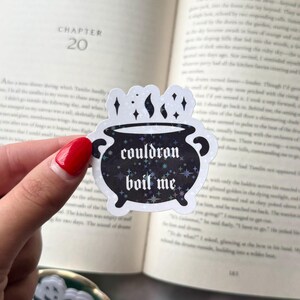 Puede incluir: Una pegatina negra con un diseño de caldero y las palabras "cauldron boil me" en una fuente estilizada. La pegatina tiene un efecto holográfico y brillante con estrellas y destellos. La pegatina se sostiene frente a un libro abierto.