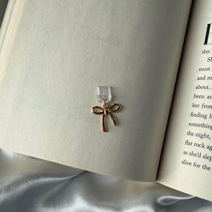 May include: A gold-colored bow-shaped earring with a clear backing is displayed against the backdrop of an open book. The earring's delicate design and the book's pages create a soft, elegant aesthetic. The earring is a jewelry item.