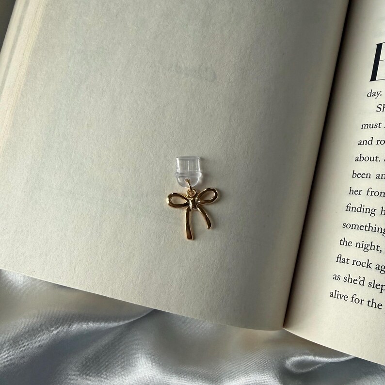 May include: A gold-colored bow-shaped earring with a clear backing is displayed against the backdrop of an open book. The earring's delicate design and the book's pages create a soft, elegant aesthetic. The earring is a jewelry item.