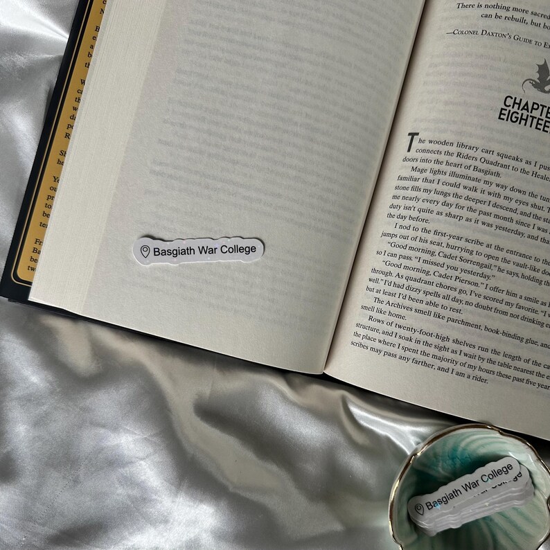 May include: An open book with a white bookmark that reads "Basgiath War College." The book is on a silky, light-colored surface. A small, decorative dish with a matching label is in the foreground.