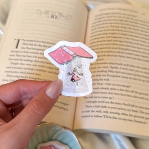 May include: A holographic sticker featuring a pink book with stars and a woman in a pink dress. The text "TAKE ME AWAY" is visible. The sticker is held in front of an open book.