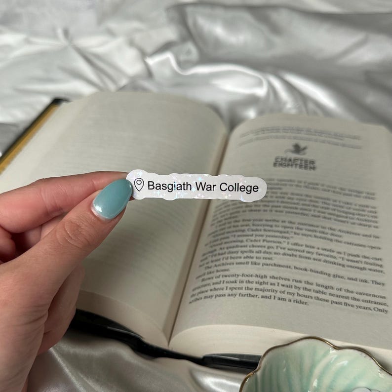 May include: A rectangular sticker with the text "Basgiath War College" in black. The sticker is held over an open book with text. The background is a white, silky fabric. The sticker has a location pin icon.