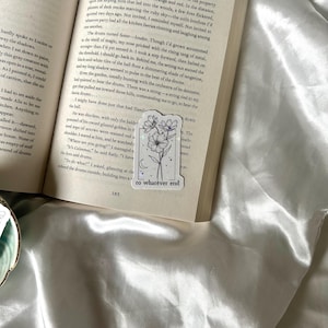 May include: A white sticker with a floral design and the text "to whatever end" is placed on an open book. The sticker features a black line drawing of flowers and a crescent moon. The book is on a white satin surface.