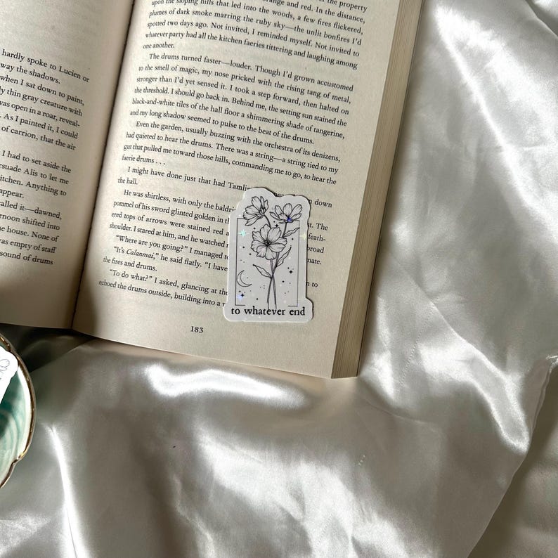 May include: A white sticker with a floral design and the text "to whatever end" is placed on an open book. The sticker features a black line drawing of flowers and a crescent moon. The book is on a white satin surface.