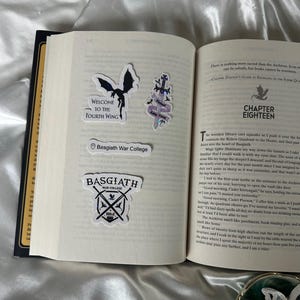 Peut inclure: Livre ouvert avec plusieurs autocollants. Les autocollants comprennent un dragon noir, une &eacute;p&eacute;e avec des accents violets et du texte qui dit "Welcome to the Fourth Wing", "Basgiath War College" et "Basgiath War College" avec des &eacute;p&eacute;es crois&eacute;es.