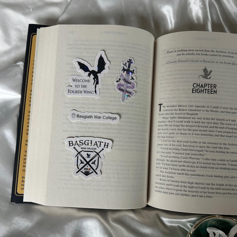 Peut inclure: Livre ouvert avec plusieurs autocollants. Les autocollants comprennent un dragon noir, une &eacute;p&eacute;e avec des accents violets et du texte qui dit "Welcome to the Fourth Wing", "Basgiath War College" et "Basgiath War College" avec des &eacute;p&eacute;es crois&eacute;es.