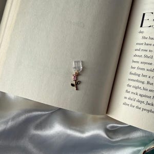 May include: A clear, rectangular bookmark with a gold-colored rose charm. The charm features a pink rose with green leaves. The bookmark is placed in an open book with text visible on the pages.