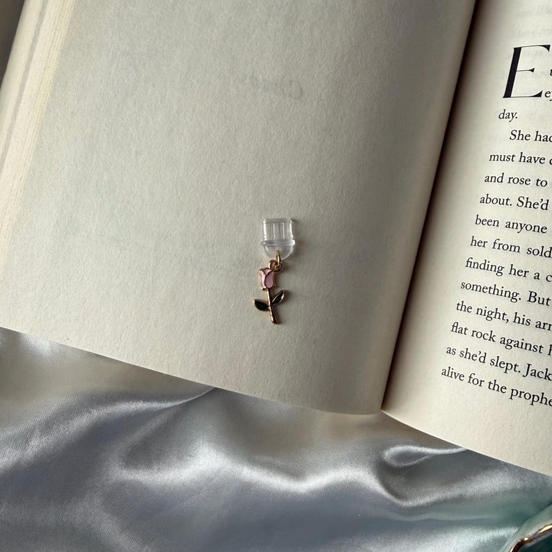 May include: A clear, rectangular bookmark with a gold-colored rose charm. The charm features a pink rose with green leaves. The bookmark is placed in an open book with text visible on the pages.