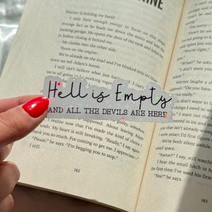 Puede incluir: Un marcapáginas transparente y brillante con el texto "Hell is Empty AND ALL THE DEVILS ARE HERE" en escritura negra. El marcapáginas se sostiene sobre un libro abierto, con el texto del libro visible en el fondo.