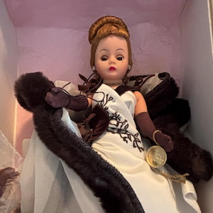 May include: A porcelain doll wearing a white gown with brown fur trim and a brown fur stole. The doll has brown hair styled in a bun and is wearing a pearl necklace and earrings. The doll is in a white box with pink lining.