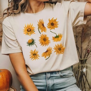 Può includere: Una maglietta color crema con un motivo di girasoli gialli. La maglietta è indossata da una persona con capelli lunghi e biondi e jeans blu.